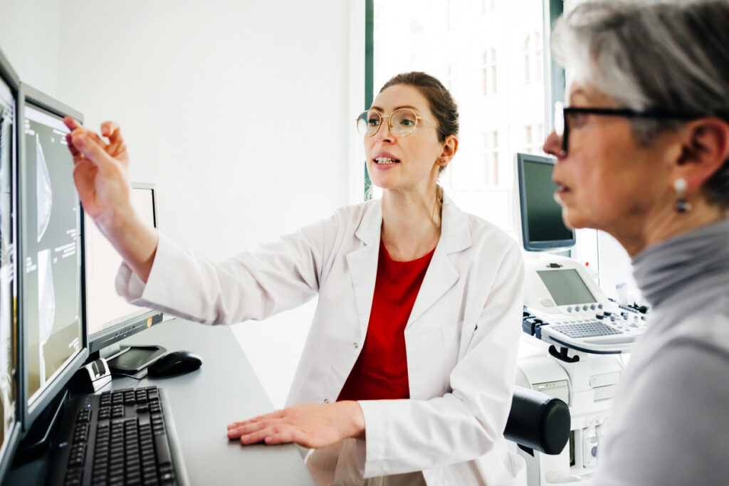 A young female doctor points to a computer monitor with CT scans while explaining test results to a senior female patient seated beside her.